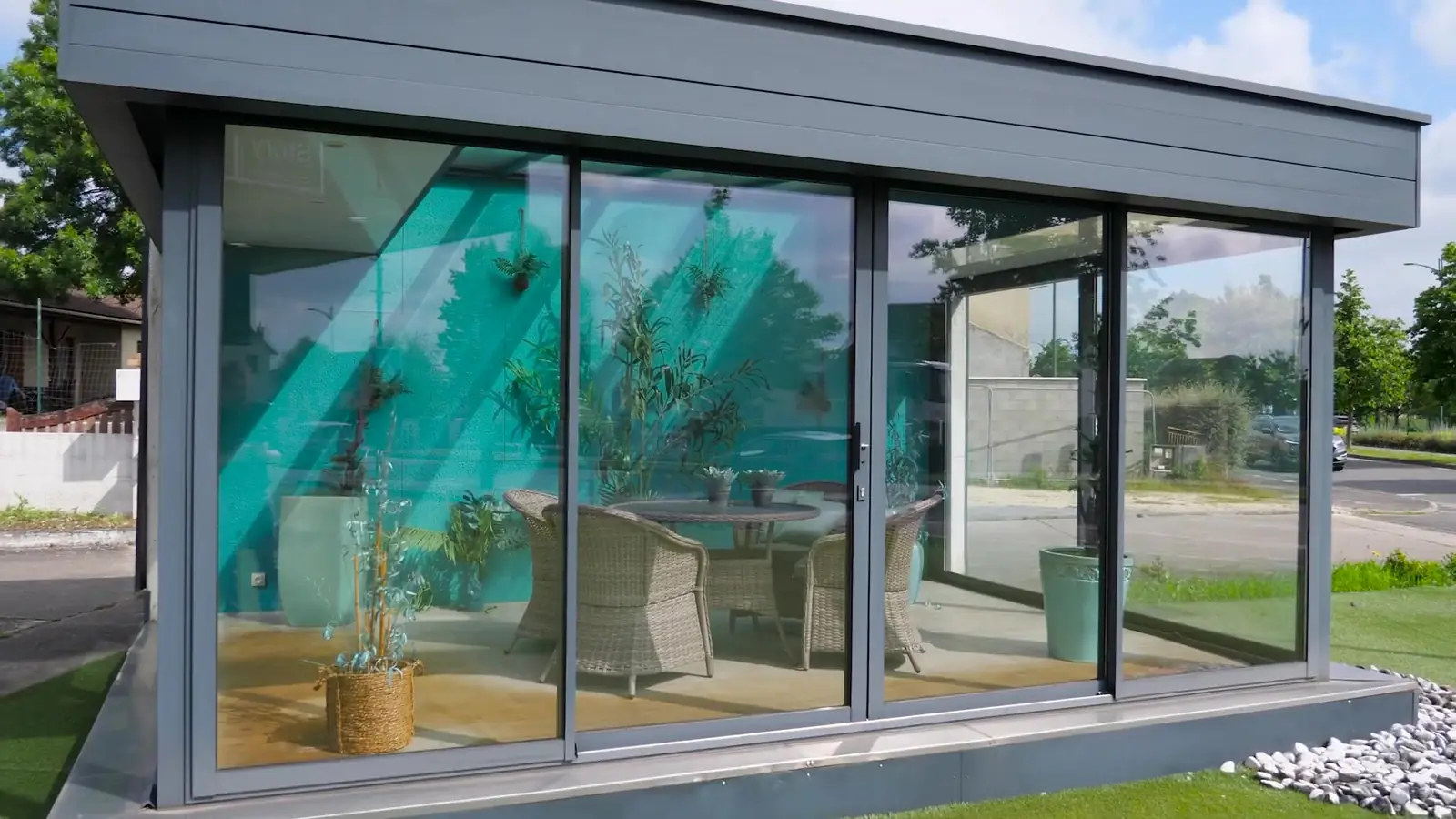 Véranda à toit plat, baies vitrées et structure grise, meublée de fauteuils en résine tressée